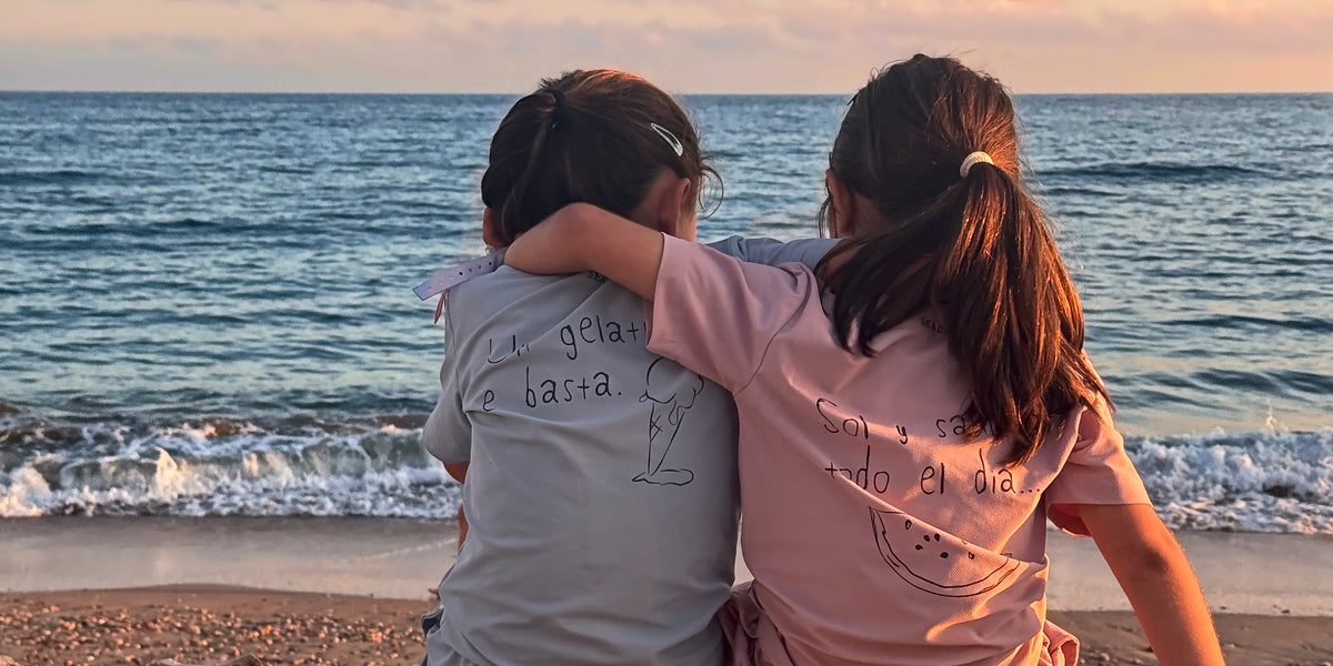 Two girls sitting side by side on a Mediterranean beach wall at sunset, arm in arm, in Seabass Kids UV T-Shirts Summer Flavours in light blue and pink.